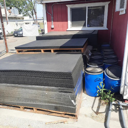 Stacks of black mats and blue barrels are arranged outside a red building. The scene is industrial and organized, with clear blue skies above.